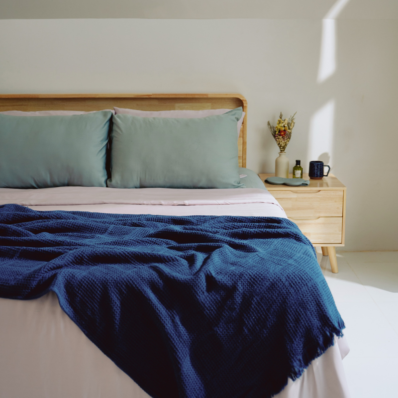 Bed with purple bedsheets, blue throw blanket and green pillows in a bedroom setting.
